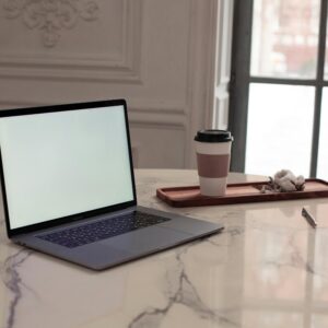 A minimalist interior setup featuring a laptop, coffee cup, and pen on a marble table by a window.