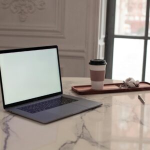 A minimalist interior setup featuring a laptop, coffee cup, and pen on a marble table by a window.